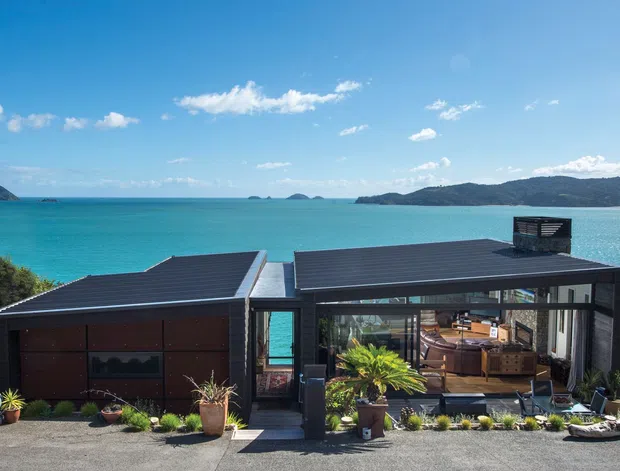 Contemporary single-storey coastal home viewed from the exterior, with dark metal cladding and low-pitched roofs. Large glazed living spaces open toward a turquoise bay, with a garage-like wing, landscaped entry planting, and a rooftop chimney or ventilation structure. Distant hills and islands form the backdrop.