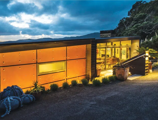 A compact modern coastal building viewed from outside at dusk, set against a steep vegetated hillside and expansive water view. The facade combines dark horizontal cladding with large warm orange-brown metal panels, while illuminated glazing reveals a warmly lit interior. A gravel approach, planted succulents, timber steps and railing complete the scene.