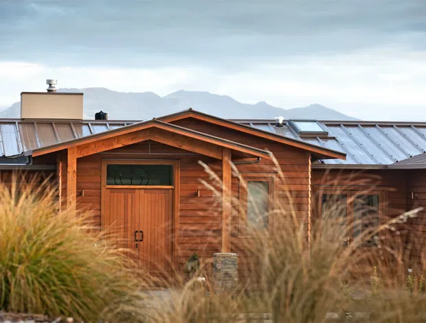 A rustic single-storey timber-clad building with a prominent gabled entrance porch and warm-toned wooden door. Corrugated metal roofing, roof skylights, and a chimney are visible, with ornamental grasses in the foreground and mountain ranges in the background. The image emphasizes the exterior facade and natural setting.