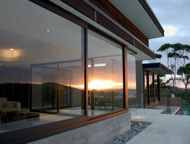 Exterior architectural view of a contemporary, multi-level residence at sunset. Large floor-to-ceiling glazed openings are framed in dark timber or metal and set above a raw concrete base, with horizontal timber cladding and projecting flat roof planes. A pale paved terrace, smooth river stones, glass balustrades, and a glimpse of a pool surround the building. Interior living areas and outdoor seating are visible through the glazing, with reflected sunset light creating a dramatic atmosphere.