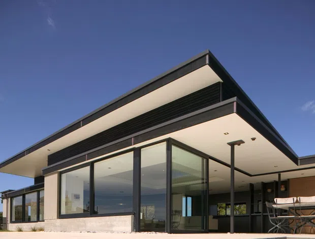 Exterior view of a contemporary single-storey residence with strong horizontal roof planes, deep cantilevered eaves, dark metal framing, extensive floor-to-ceiling glazing, and an open covered outdoor dining area. The restrained black, white, and concrete palette creates a crisp modern architectural character.