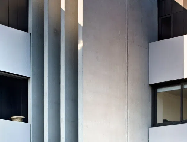 Close-up exterior view of a contemporary multi-storey building with a strong geometric facade. Tall pale concrete or rendered vertical fins project from a broad central wall, framed by dark charcoal cladding, black-framed glazing, and white balcony parapets. Tropical planting is visible at the base beneath a clear blue sky.
