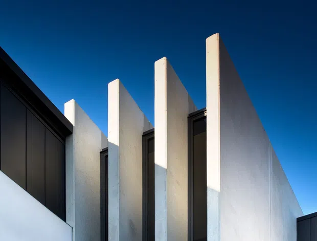 Close-up exterior architectural view showing a contemporary facade composed of tall, projecting white rendered fins or blade walls set alongside narrow dark-framed vertical windows. A dark roof or wall volume intersects the composition at left and right, contrasted against a clear deep blue sky. The image emphasizes strong vertical repetition, geometric massing, shadow, and minimalist material contrast.