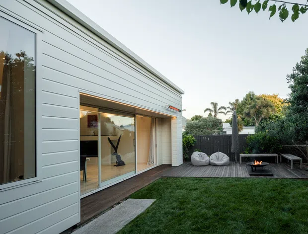 Exterior view of a contemporary single-storey home with pale horizontal weatherboard cladding, tall narrow glazing, and a large sliding glass opening onto a timber deck. The deck leads to a landscaped backyard with outdoor seating, a small fire feature, fencing, and mature planting.