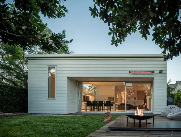 A compact modern single-storey pavilion or extension set in a leafy garden. The exterior is clad in pale horizontal weatherboards with a simple flat roof and large glazed sliding doors opening to a furnished interior. A raised timber deck with a low outdoor fire bowl occupies the foreground, while mature trees frame the scene.