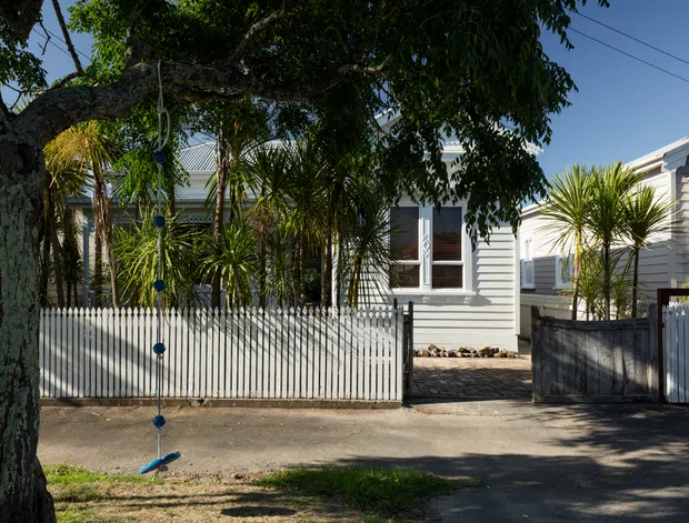 Street-facing view of a compact light-grey weatherboard house behind a white picket fence, with a paved entry path, low front boundary walls, mature trees, and tropical-style planting. The single-storey facade features a pitched metal roof, white-framed windows, and a shaded verandah-like entry area.