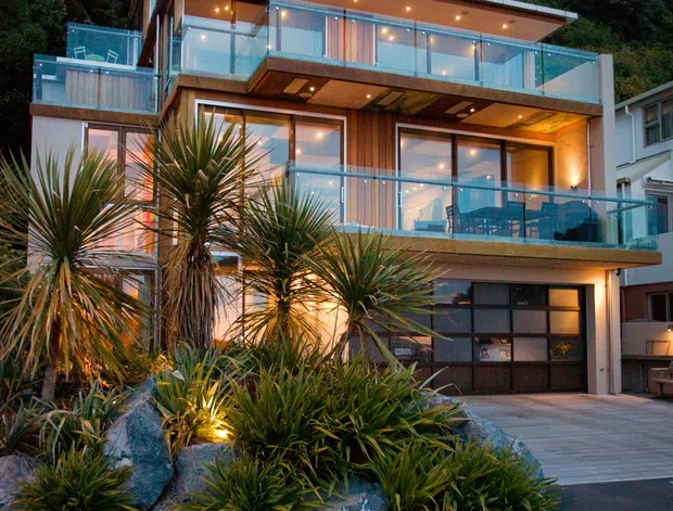 Twilight exterior view of a multi-storey contemporary residence built into a vegetated hillside. The home features broad floor-to-ceiling glazing, multiple projecting balconies with clear glass balustrades, warm timber wall cladding and soffits, recessed lighting, and a garage at ground level. Native-style spiky planting and boulders form a landscaped frontage beside the driveway and road.