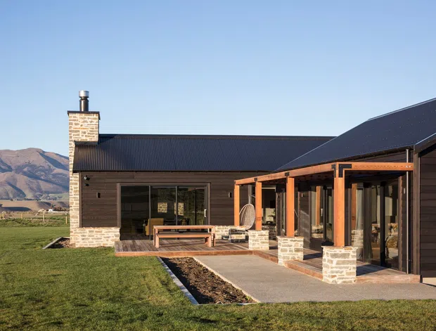 A low-slung modern rural house viewed from the exterior, with dark horizontal cladding, black metal pitched roofs, extensive glazing, and a covered courtyard terrace supported by timber posts on stone-clad bases. A brick chimney and mountain landscape are visible in the background.