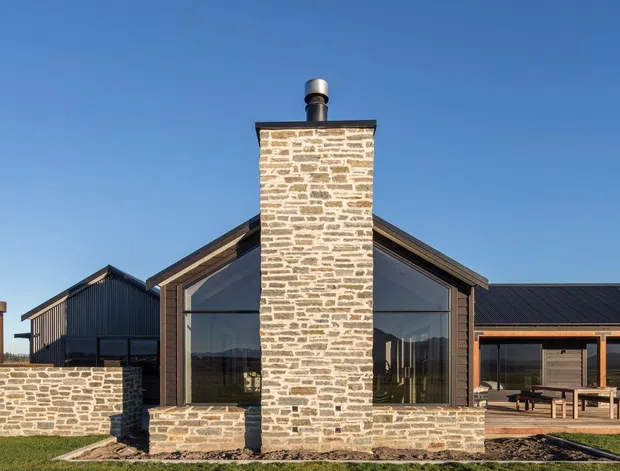Exterior photograph of a low-rise contemporary rural-style house under a clear blue sky. The central gabled volume features a prominent rough stone chimney and matching stone base walls, large floor-to-ceiling glazing, dark metal roofing, and dark timber or metal framing. A connected wing and separate outbuilding extend to either side, with a paved outdoor dining area visible on the right and a broad lawn in the foreground.