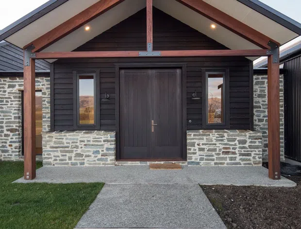 Front exterior view of a compact gable-roofed building. The facade combines dark horizontal timber cladding, grey natural-stone masonry at the lower walls, and exposed reddish timber posts and beams beneath a broad sheltered gable. A dark timber entry door is flanked by narrow windows, with a concrete path leading to the entrance and simple lawn and garden beds in front.