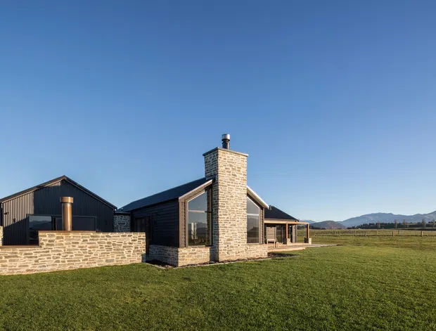 Single-storey rural residence photographed from the exterior, composed of dark vertical-clad wings and prominent natural stone masonry walls and chimney. The home has pitched gable roofs, large glazed openings, a low stone courtyard wall, and sits in an expansive grassy landscape with distant mountains under a clear sky.