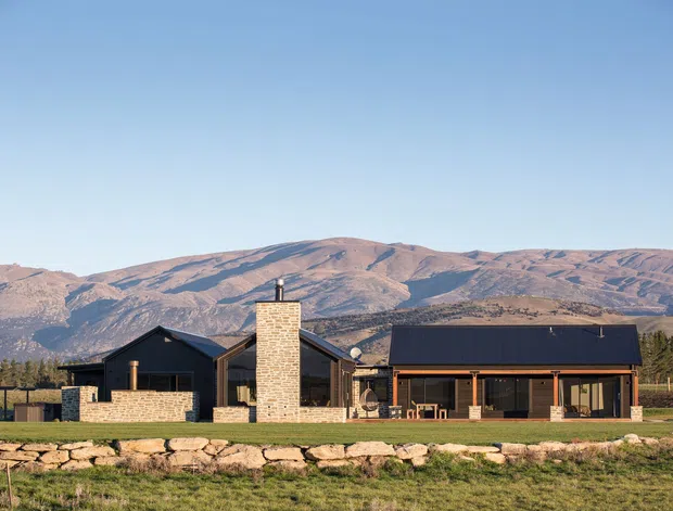 A low, elongated contemporary rural residence set in an open landscape, with dark charcoal cladding and roofing, extensive glazing, and a prominent pale brick chimney. A stone retaining wall runs across the foreground, while expansive mountain ranges form the distant backdrop under a clear blue sky.