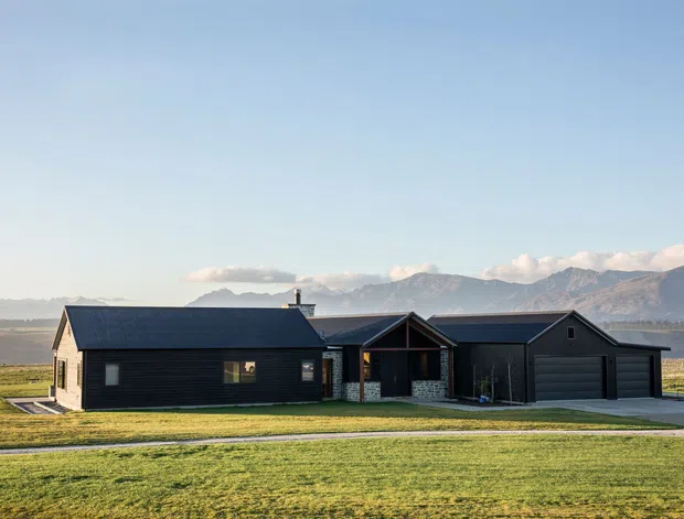 A single-storey rural home with dark horizontal cladding, multiple pitched gable roofs, and a central covered entry. The house sits in an expansive open grassy landscape with dramatic mountain ranges visible in the background. A broad driveway leads to an attached double garage, and the exterior composition has a simple contemporary lodge or farmhouse character.