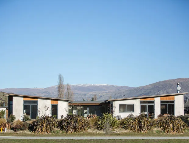Single-storey contemporary residential building viewed from the front, composed of low horizontal wings with pale rendered walls, dark-framed glazing, warm timber fascia accents, and a central recessed connection. Native-style landscaping occupies the foreground, with dry mountain terrain and a clear blue sky in the background.