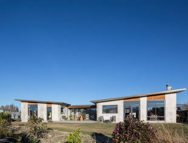 Single-storey contemporary residential house viewed from the front, composed of low horizontal wings with broad overhanging flat or very low-pitched roofs, pale rendered walls, warm timber cladding accents, and extensive dark-framed glazing. The building sits within a landscaped garden with ornamental planting and a visible covered connection between the main volumes. Clear blue sky and surrounding vegetation form the background.