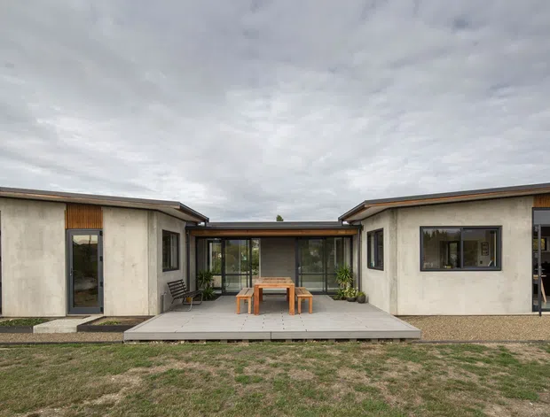 Single-storey contemporary residence viewed from the front courtyard. The low, horizontally composed building has pale rendered walls, warm timber cladding accents, dark-framed glazing, and shallow pitched roof forms arranged around a central covered outdoor dining area and concrete terrace.