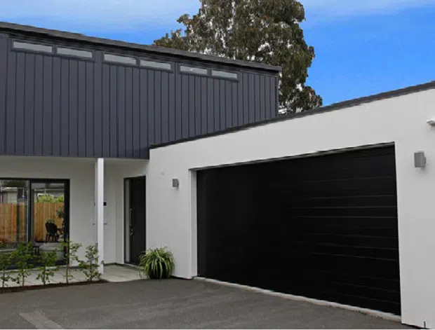 Contemporary single-storey residential exterior with a white rendered facade, black horizontal garage door, and a contrasting dark vertical-panel upper volume. The home includes a recessed entry, large window, small landscaped planting, and a paved driveway under a clear blue sky.