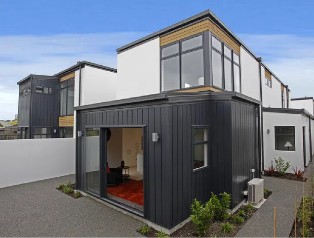 Contemporary multi-level residential building with crisp white rendered walls, dark vertical metal cladding, timber-toned trim, and extensive upper-level glazing. The image is taken from the exterior, showing a paved entry path, landscaped planting beds, and a partially visible interior through the open glazed door.