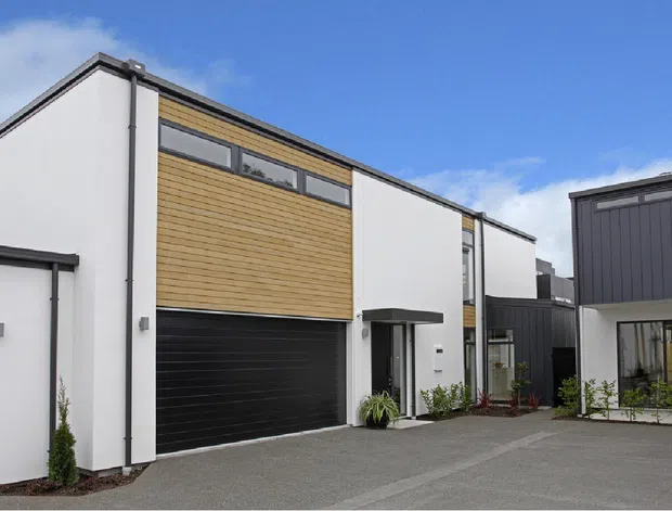 Exterior view of a contemporary multi-storey residential development with clean white rendered walls, black garage doors and trim, warm horizontal timber cladding, narrow clerestory windows, and dark vertical cladding on the adjoining building. A paved shared driveway and landscaped planting sit in front of the homes under a bright blue sky.