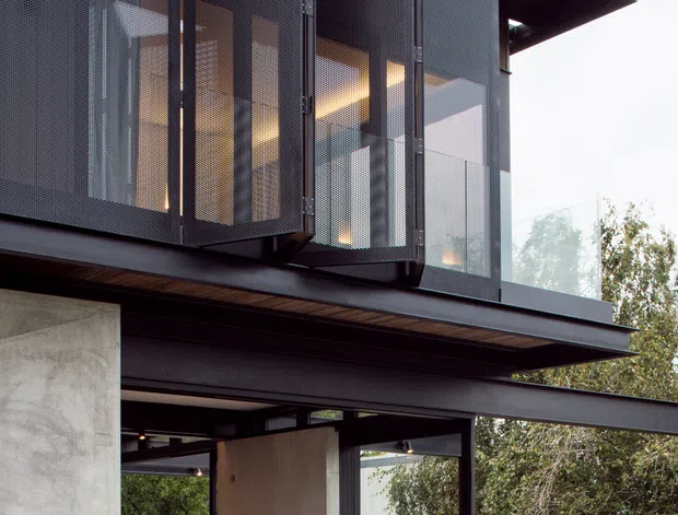 Two-storey modern residence with a dark steel-framed facade, projecting upper-level perforated metal privacy screens, extensive glazing, exposed structural beams, and a concrete lower-level terrace or living area. The architecture combines crisp horizontal roof planes, raw concrete walls, timber soffit accents, and a strong indoor-outdoor connection to surrounding trees.