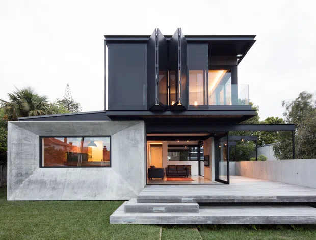 Contemporary two-storey residence with a dramatic dark metal upper volume, projecting roof planes, large glazed openings, and a raw concrete ground-floor pavilion. A broad concrete terrace and stepped platform lead to an open living area, with landscaped garden edges visible around the house.