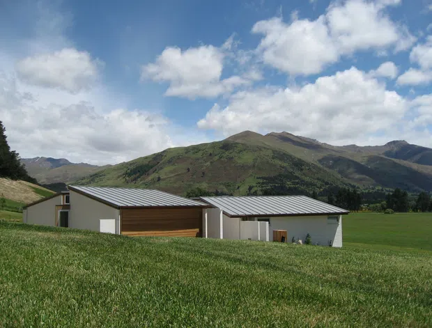 A low-profile contemporary rural building complex set in a green mountain valley. The buildings have simple pale rendered walls, broad corrugated metal roofs, and a prominent warm timber-clad garage or entrance volume. Open pasture dominates the foreground, with steep wooded and grassy mountains forming the backdrop under a partly cloudy sky.
