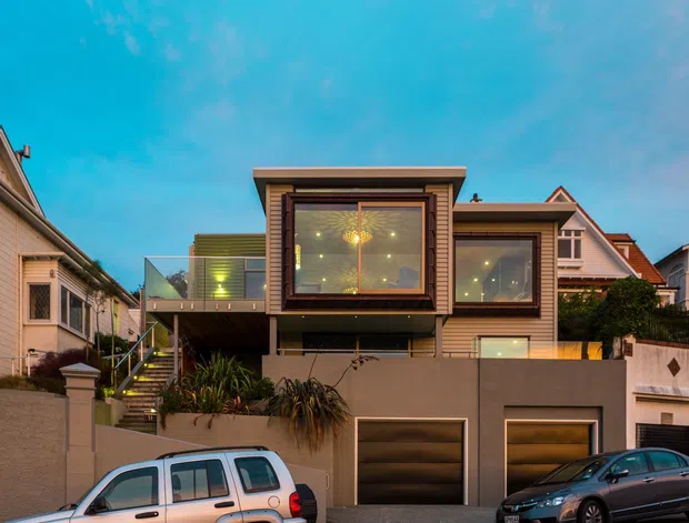 Street-facing exterior view of a contemporary multi-level residence on a sloping urban site. The home features projecting box-like upper volumes with extensive glazing, horizontal timber-toned cladding, recessed downlighting, balconies, and an integrated lower-level double garage. Neighbouring houses, retaining walls, landscaping, and parked vehicles are visible in the foreground and surroundings.