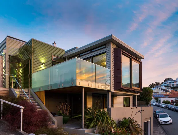 Exterior view of a contemporary multi-level urban residence on a sloping site, featuring articulated horizontal cladding, projecting upper volumes, extensive floor-to-ceiling glazing, and a translucent glass-balustraded terrace. A stair leads up through landscaped planting, with neighbouring homes and a street visible beyond. The image is photographed at dusk with warm interior lighting and a blue-pink sky.