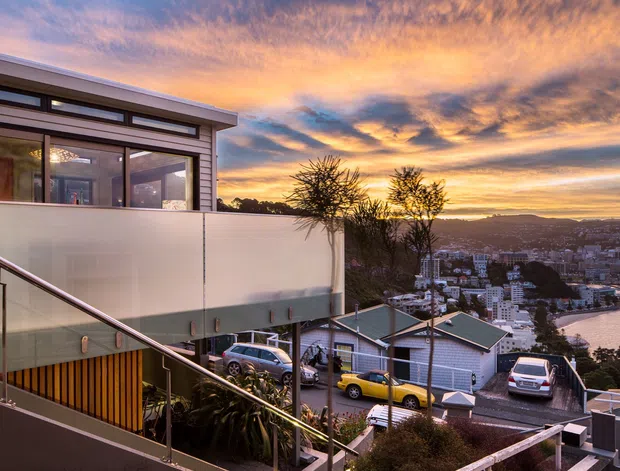 An elevated exterior view of a contemporary multi-level residential house on a steep urban hillside at sunset. The image shows a flat-roofed upper volume with extensive glazing, translucent privacy screening, timber-toned vertical cladding, metal handrails, and stepped outdoor circulation. A dense coastal city and inlet are visible beyond the property.