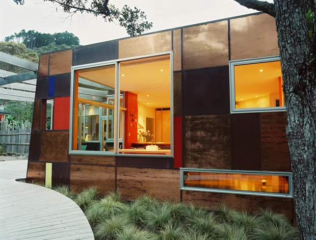 Exterior view of a compact modern dwelling set among mature trees and ornamental grasses. The facade is clad in weathered rust-brown metal panels with dark framed windows and large glazed openings revealing a warmly lit interior. A curved pale timber boardwalk leads alongside the building, while a long low glazed opening forms a distinctive horizontal feature near the base.