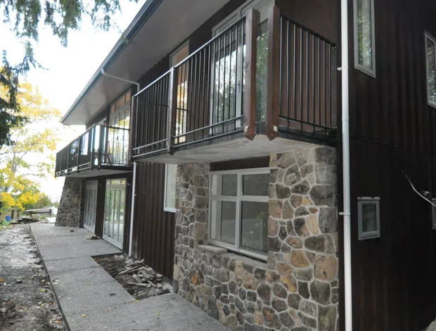 Exterior view of a multi-level residential building with dark vertical timber cladding, exposed natural stone masonry at the lower walls, grey metal balcony railings, white-framed windows and doors, and a sloped roof with broad eaves. A narrow concrete walkway runs alongside the building.