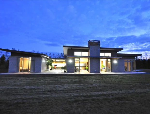 Wide dusk exterior view of a low, single-storey contemporary residence with a strongly horizontal flat roof, projecting roof planes, extensive glazed openings, and a central taller masonry-clad volume with a screened upper section. Interior lighting glows through the windows, while the home sits behind an open lawn and dark tree line.
