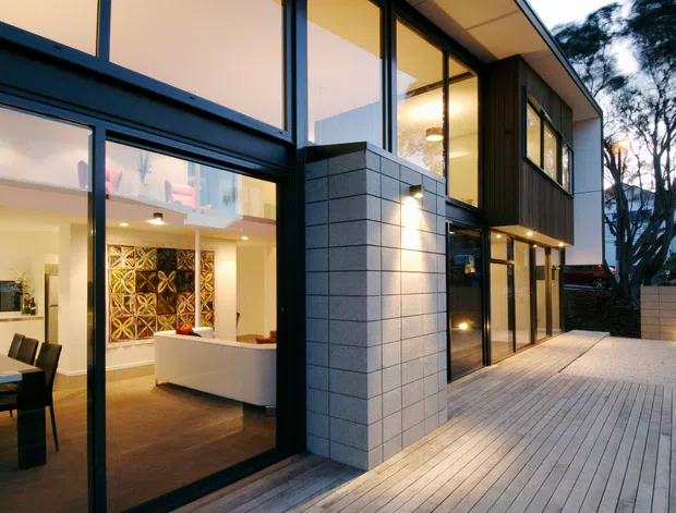 Contemporary multi-level residential exterior viewed from a timber deck at dusk, with extensive floor-to-ceiling glazing, dark window frames, a pale masonry feature wall, and dark vertical cladding. The illuminated interior visibly includes an open-plan kitchen, dining area, and living room, while mature trees and a neighbouring property form the background.