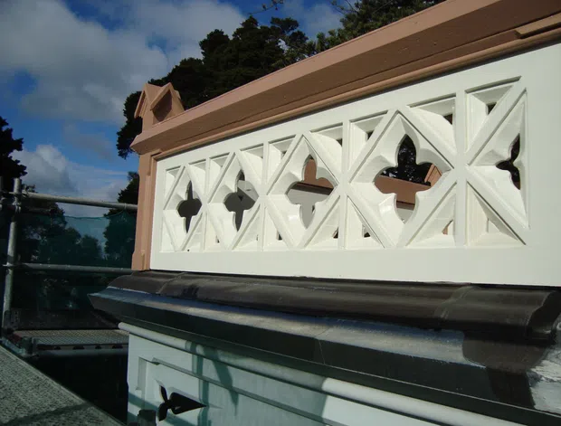 Close-up exterior view of an ornate white geometric breeze-block balustrade or parapet, set within a pale pink rendered frame above dark roofing or trim. The decorative screen features repeated angular diamond and pointed-arch openings, with a small projecting gable-like detail at the upper left. Trees and sky are visible in the background.