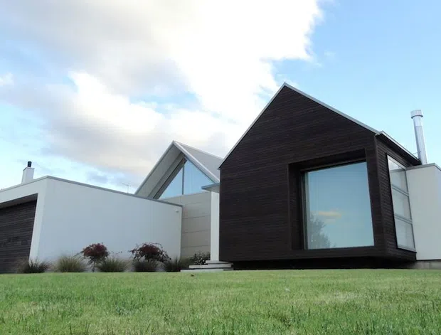 Contemporary single-storey residential house set in a broad lawn, composed of crisp white rendered volumes and a prominent dark timber-clad gabled pavilion with large glazing. The image shows a sculptural, asymmetrical roof composition, minimal detailing, landscaped grass, and a mountainous backdrop.