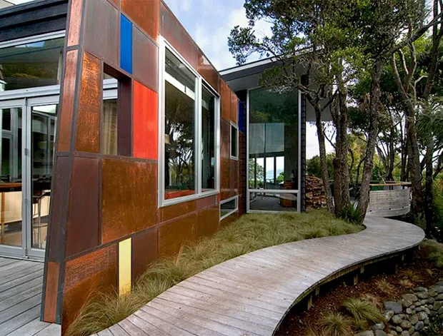 Exterior view of a contemporary, low-profile building set among mature trees and landscaped planting. The facade combines large reflective glazing with weathered rust-coloured metal panels, including blue and red accent panels. A curved timber boardwalk leads around the building, bordered by ornamental grasses and dark stone landscaping. Interior spaces are visible through the glazing.