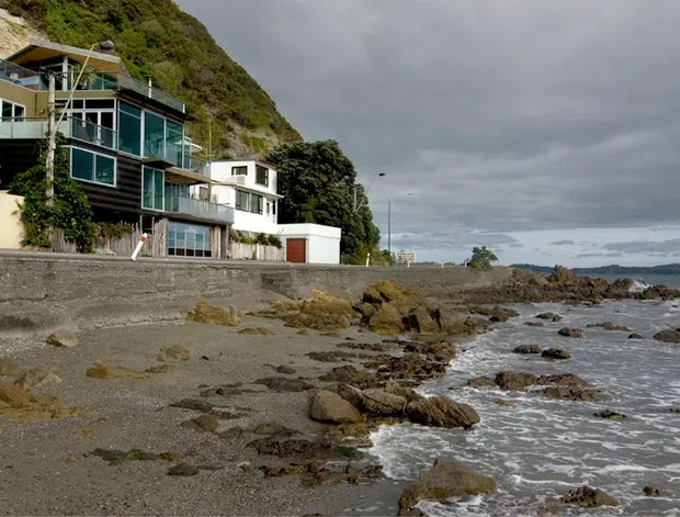A modern multi-level coastal house built directly along a rocky shoreline, with extensive glazed living spaces, projecting balconies, dark timber cladding, and pale rendered walls. A steep vegetated hillside rises behind the home, while a seawall, rocky beach, and calm water extend across the foreground and right side. The overcast atmosphere gives the scene a cool, dramatic coastal character.