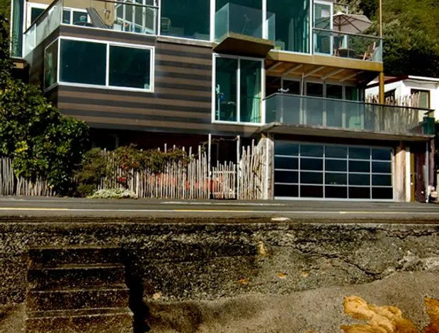 A multi-level contemporary house built into a densely vegetated coastal hillside, viewed from across exposed rocky shoreline. The home features extensive floor-to-ceiling glazing, dark horizontal cladding, multiple glass-balustraded terraces and balconies, a flat projecting roof structure, and an integrated garage at street level.