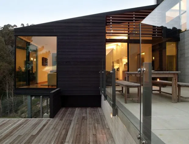 Contemporary multi-level house set among trees, viewed from an elevated timber deck. The dark horizontal-clad facade combines projecting glazed volumes, expansive glass doors, a slatted timber privacy screen, and a bright outdoor dining terrace with a long timber table and benches. Warm interior lighting contrasts with the dark exterior cladding and surrounding bushland.