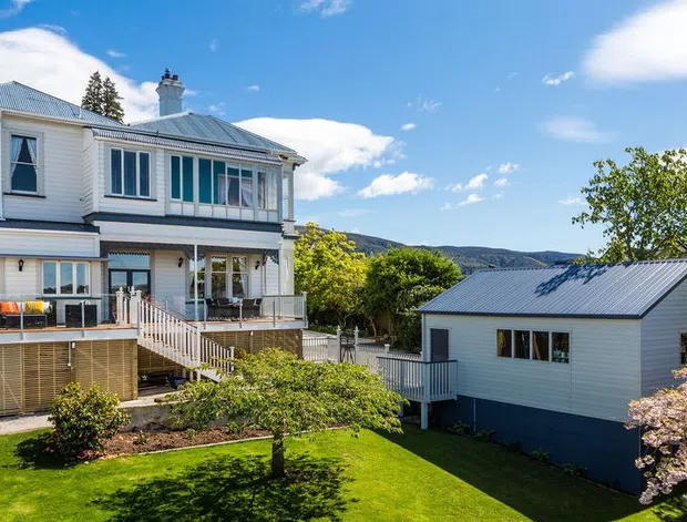 Exterior view of a white weatherboard two-storey house with a metal hipped roof, verandah, balcony, exterior stairs and a separate matching outbuilding. The landscaped garden sits beside a calm bay or inlet with hills in the background under a bright blue sky.