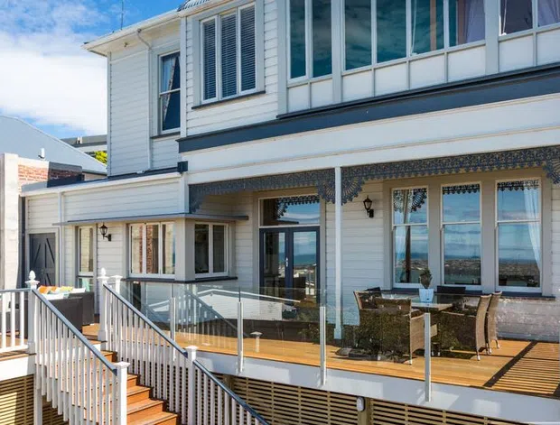 Exterior view of a multi-level coastal-style home with white weatherboard cladding, extensive glazing, decorative patterned tile detailing beneath the upper balcony, and a timber deck with glass balustrades and stairs. The architecture overlooks a bright blue coastal or waterfront setting visible through the openings.