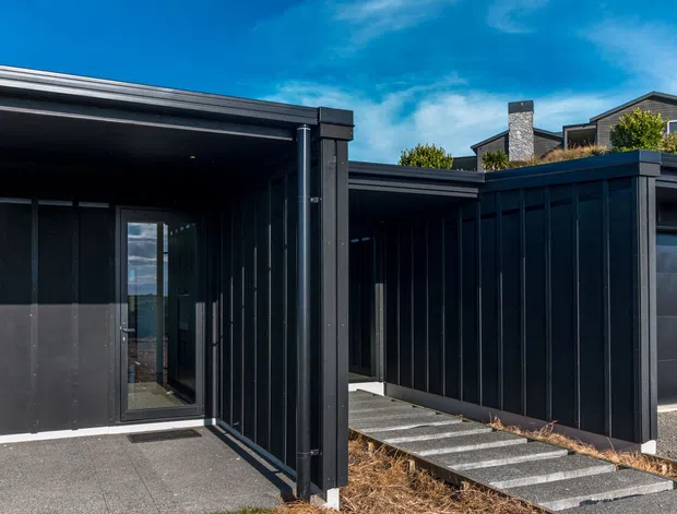 Modern dark-clad residential exterior with low-slung flat-roofed forms, recessed entry glazing, a covered walkway, and a concrete stepping-stone path. The facade combines black vertical cladding with dark garage doors and strong horizontal rooflines.