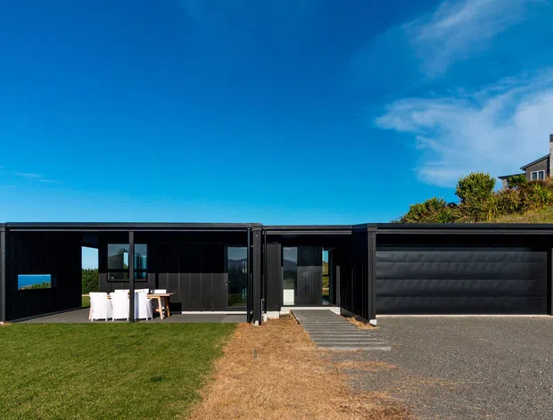 A low-profile, single-storey contemporary house with a dark black facade and flat roof, viewed from the front. The building includes a sheltered outdoor dining area, large glazing, a central entry path, and an integrated black garage door. It sits on a grassy coastal-looking site beneath a bright blue sky, with another house visible on the hillside behind.