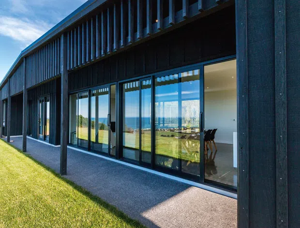 Exterior view of a long, contemporary single-storey building with dark vertical cladding, extensive floor-to-ceiling glazing, and a covered verandah or walkway supported by slender posts. The glazing reflects a broad grassy landscape and reveals a bright interior with dining furniture. The building has a strong linear form, deep roof overhang, and upper-level vertical screening or louvres.