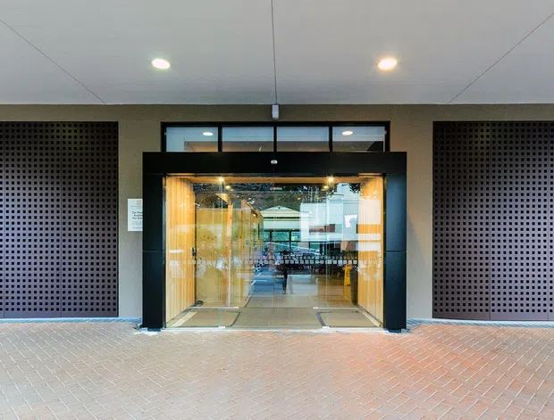A symmetrical commercial building entrance with a central glazed automatic doorway framed in black, flanked by two large dark perforated screen panels. The entrance opens into a brightly lit interior, while pale tiled paving extends across the foreground. The facade has a restrained contemporary design with recessed downlights and strong geometric alignment.