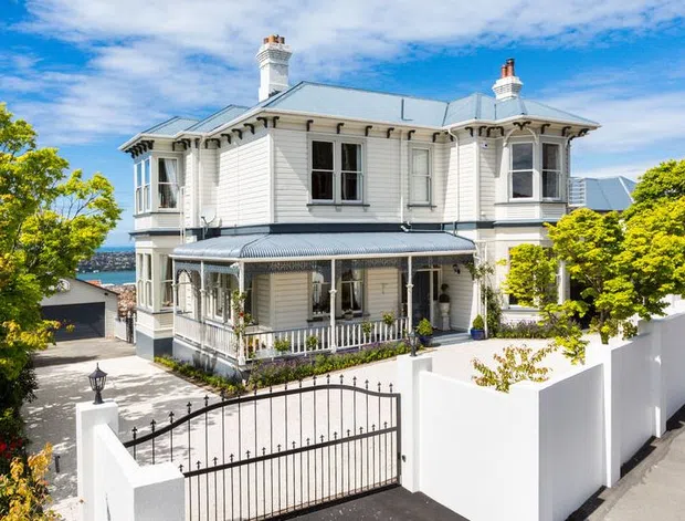 A grand two-storey historic villa with a pale weatherboard facade, ornate verandah, bay windows, decorative trims, chimneys, and a low-pitched metal roof. The property has a gated driveway, white masonry boundary wall, established trees, and a glimpse of blue water beyond the house.