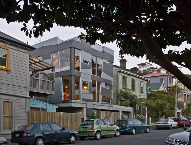 Street-level view of a contemporary multi-storey residential building inserted among older timber-clad homes in an established urban neighbourhood. The main building has a charcoal-grey rectilinear facade, extensive floor-to-ceiling glazing, projecting box forms, balconies, and a lower concrete or masonry base with timber fencing. Mature street trees and parked cars frame the scene.