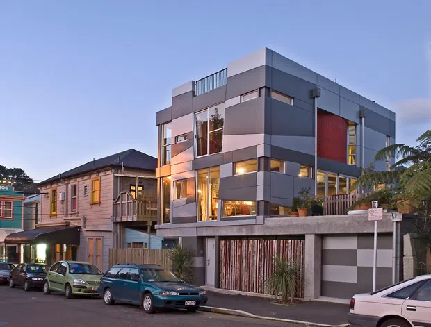 Exterior streetscape photograph of a contemporary multi-storey urban residence with a bold rectilinear form, grey panelled facade, extensive glazing, rooftop terrace railing, warm interior lighting, and a deep concrete frontage over a garage and timber slatted entry screen. Older timber homes and other dense residential buildings sit closely alongside it, creating an inner-city suburban context.
