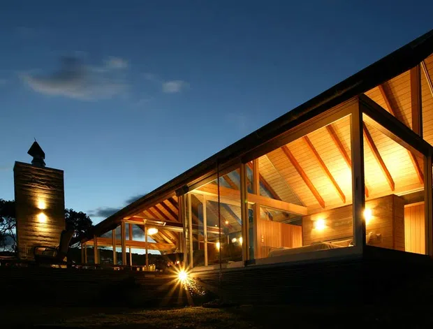 Dusk exterior view of a low-rise contemporary timber-clad building with extensive floor-to-ceiling glazing, dramatic pitched roof planes, exposed dark structural framing, and warm interior lighting. A tall illuminated chimney or sculptural masonry element stands at the left, with a covered outdoor walkway or deck extending along the facade.