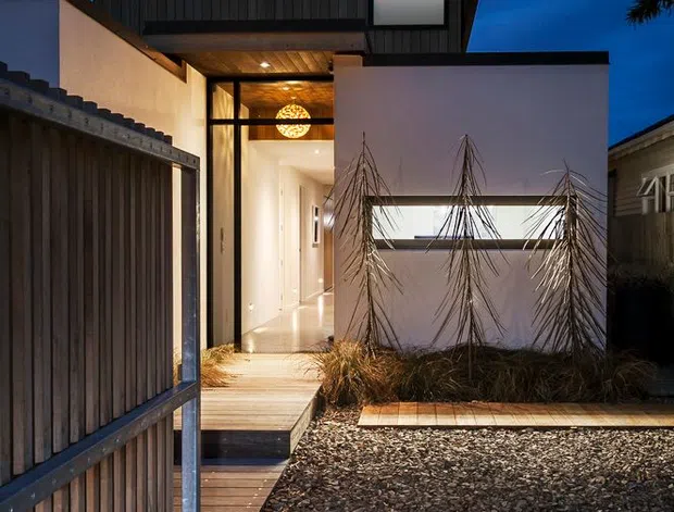 A dusk exterior view of a contemporary residential entry courtyard. A dark timber-clad, asymmetrical gabled volume sits above a brightly lit glazed entry, with white rendered walls, timber decking and walkway, gravel landscaping, and ornamental grasses. A timber and metal gate frames the foreground, while warm interior lighting contrasts with the deep blue evening sky.